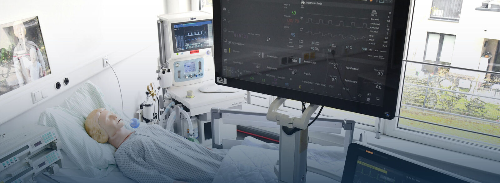 Ein medizinischer Simulationsraum mit einer Übungspuppe, die in einem Krankenhausbett liegt und Patientenbekleidung trägt. Die Puppe ist an medizinische Geräte angeschlossen, darunter Monitore, die Vitalzeichen wie Herzfrequenz und Sauerstoffsättigung anzeigen. Mehrere Bildschirme zeigen detaillierte medizinische Daten.