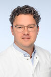 Porträtfoto eines Mannes mit dunklem Haar, Brille und weißer medizinischer Kleidung.