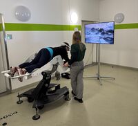 ICAROS Eine Person liegt auf einer Art Liege, trägt eine VR Brille und schaut in Richtung eines Bildschirms. Daneben steht eine Person die auch in Richtung des Bildschirms blickt.