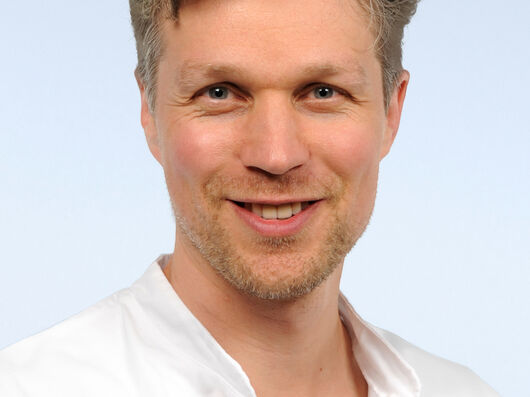 Portraitfoto von Mann mit kurzen, grauen, lockigen Haaren und Dreitagebart. Er lacht in die Kamera und trägt einen weißen Arztkittel.