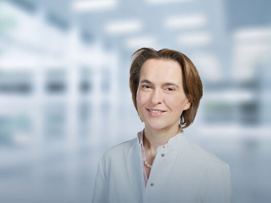 Eine Frau steht in einem modernen, gut beleuchteten Büro oder Labor. Im Hintergrund sind große Fenster und helle Lichtquellen zu erkennen, jedoch unscharf dargestellt. Die Umgebung wirkt professionell und technisch.