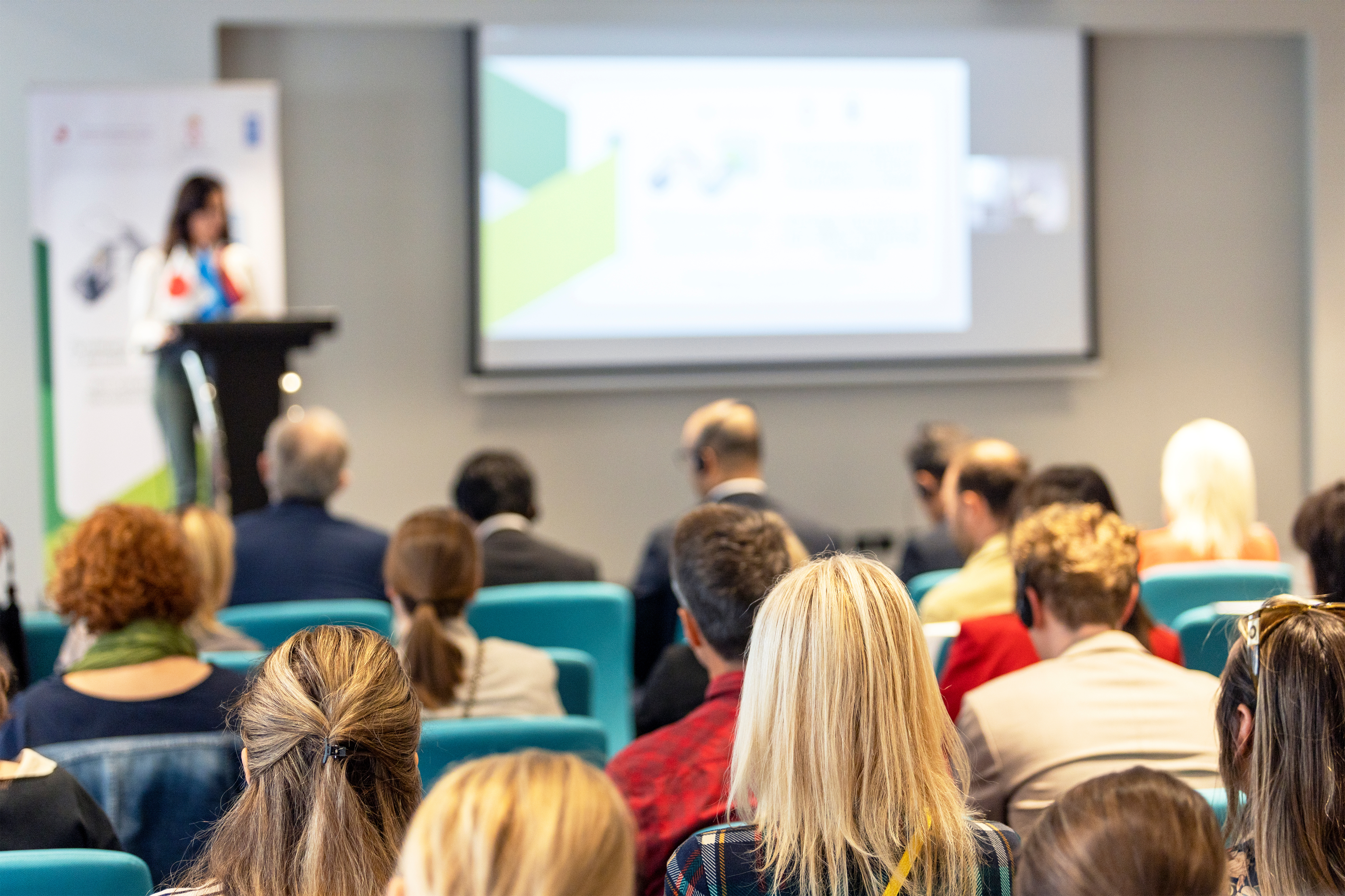 Eine Gruppe von Menschen sitzt in einem Konferenzraum und schaut auf einen Redner, der an einem Podium steht. Der Redner präsentiert Informationen, die auf einem großen Bildschirm hinter ihm angezeigt werden.