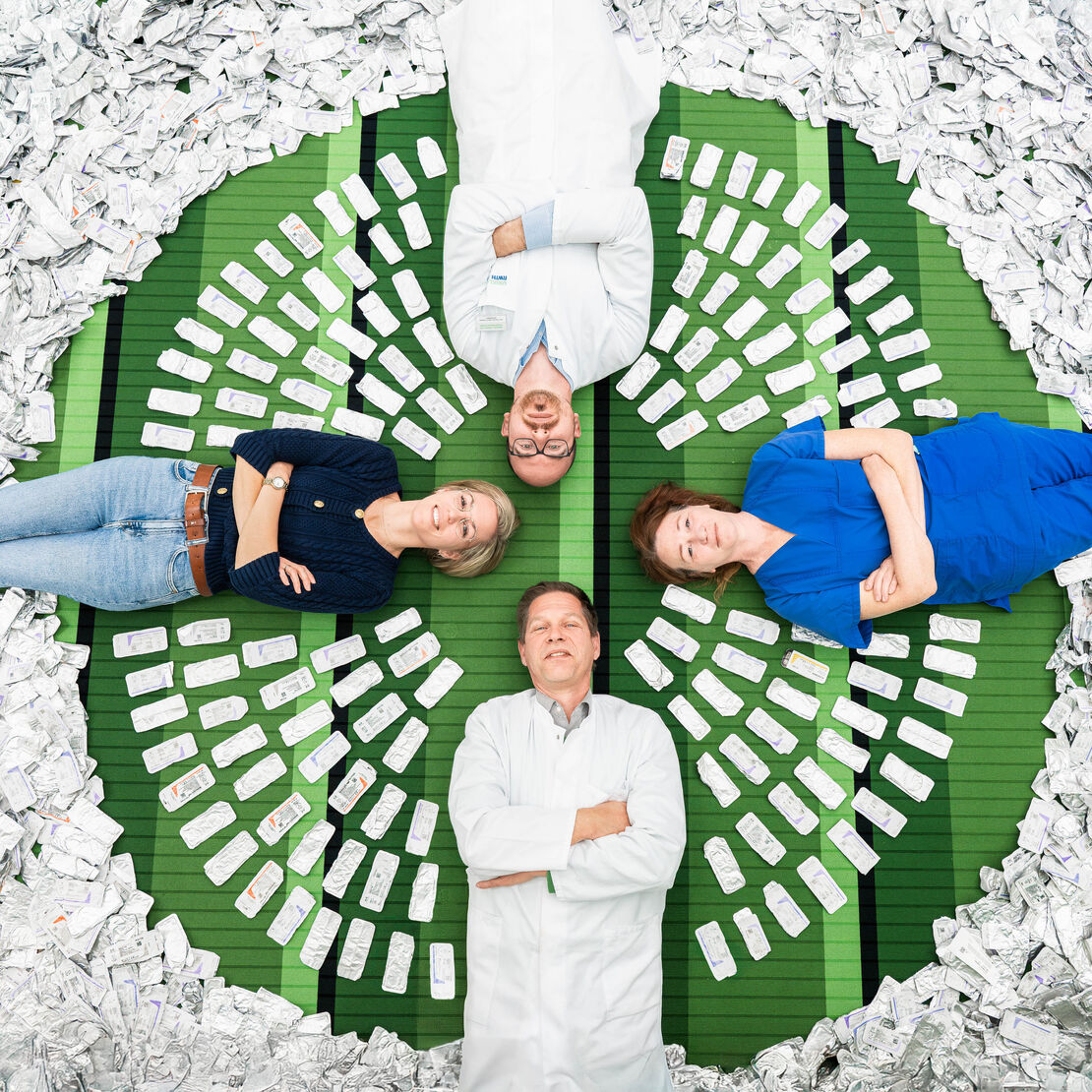 Neues Aluminium‑Recyclingprogramm im Zentral-OP Vier Personen liegen in Kreuzform auf einer grünen Fläche, umgeben von zahlreichen aluminiumhaltigen Nahtverpackungen, die kreisförmig um sie angeordnet sind.