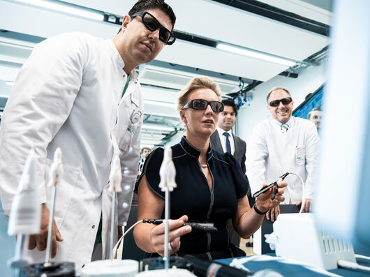 Ein Arzt im weißen Kittel mit Sonnenbrille steht neben einer Frau mit Sonnenbrille, während sie einen chirurgischen Roboter austestet