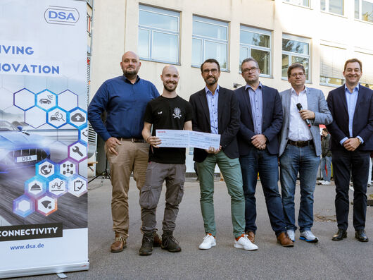 Gruppenaufnahme von sechs Männern, die freundlich in die Kamera lächeln. Zwei Männer halten einen Scheck.