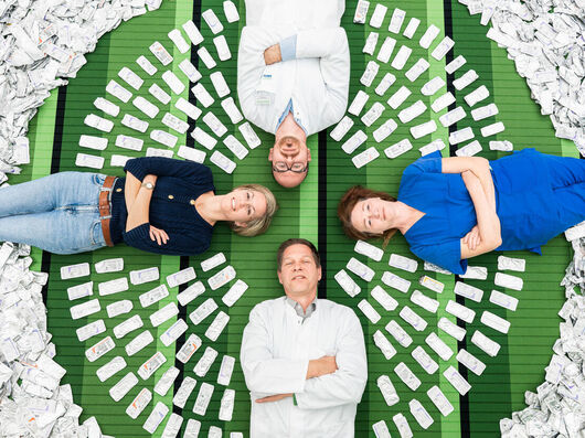 Neues Aluminium‑Recyclingprogramm im Zentral-OP Vier Personen liegen in Kreuzform auf einer grünen Fläche, umgeben von zahlreichen aluminiumhaltigen Nahtverpackungen, die kreisförmig um sie angeordnet sind.