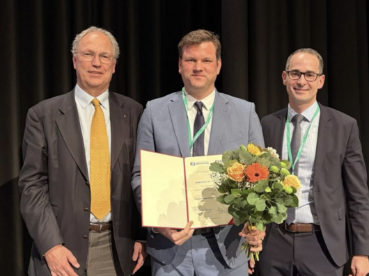 Gruppenfoto dreier Männer mittleren Alters in Anzügen. Der Mann in der Mitte hält eine Urkunde und einen Blumenstrauß.