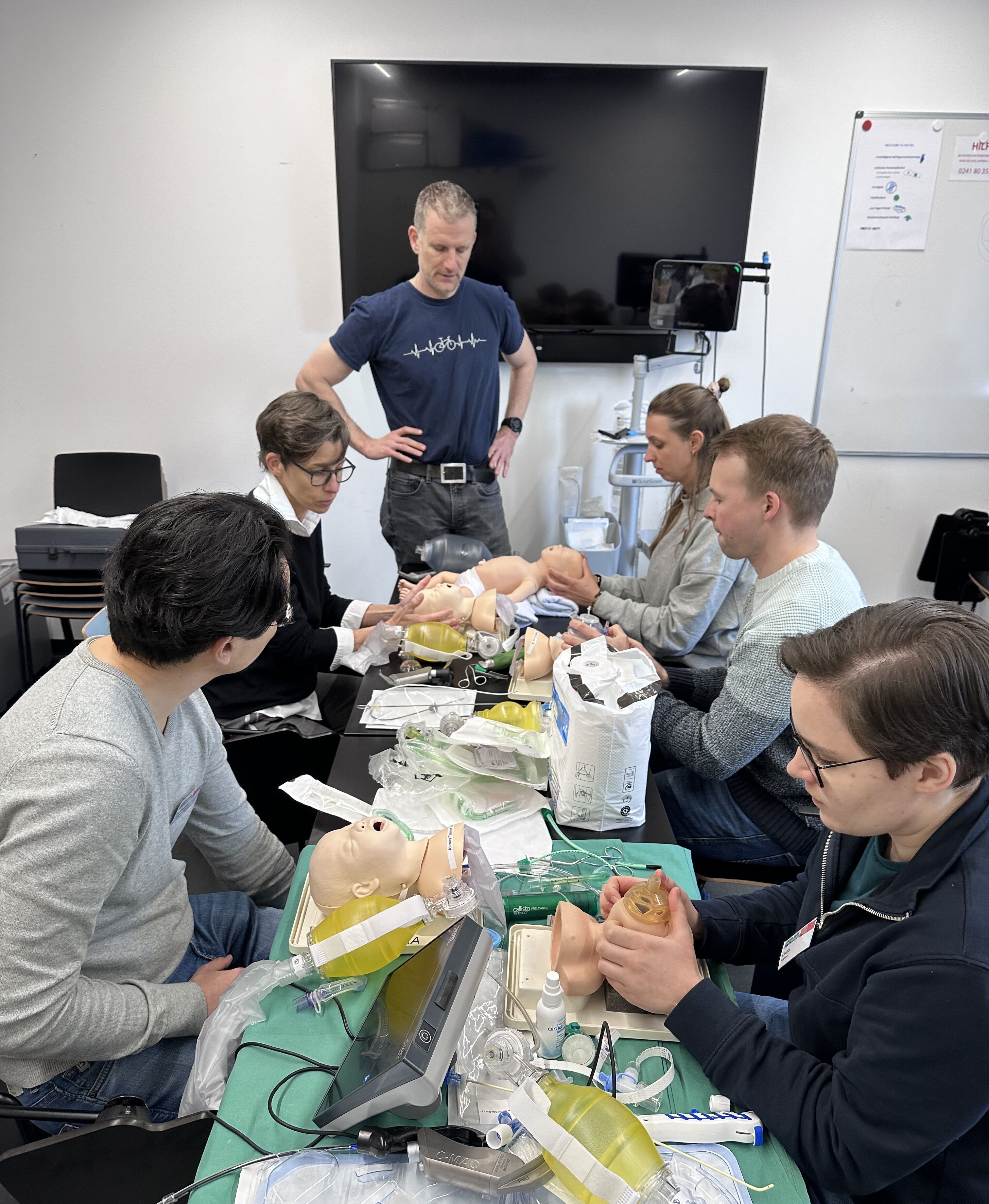 Mehrere Personen stehen um einen Tisch mit medizinischem Trainingsmaterial, darunter Säuglingspuppen und verschiedene medizinische Utensilien. Die Gruppe scheint an einer praktischen Schulung teilzunehmen und medizinische Verfahren an den Puppen zu üben