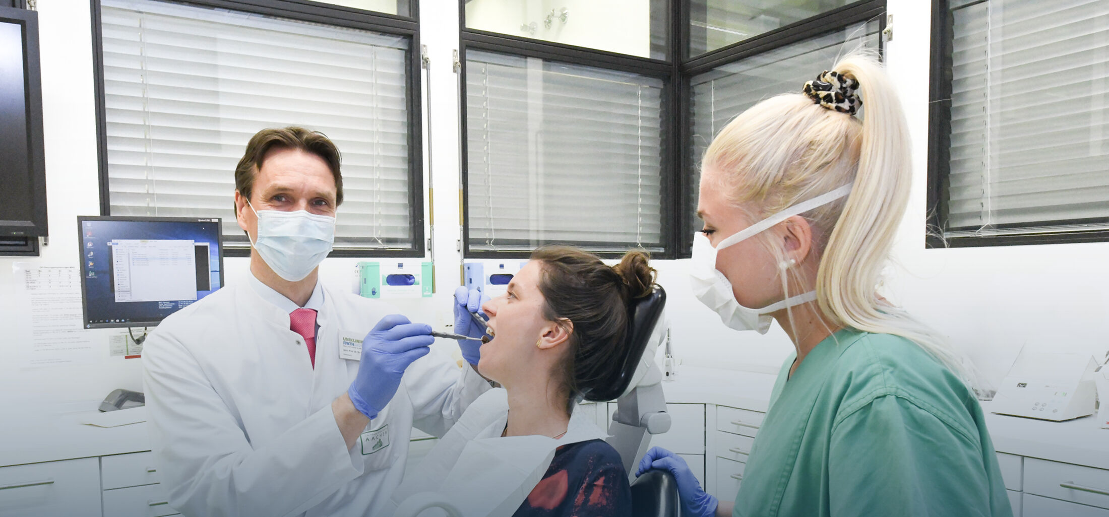 Arzt mit Maske in weißer Kleidung mit medizinischem Werkzeug in Mund von Patientin, begleitet von einer blonden Frau mit Pferdeschwanz, Maske und grüner medizinischer Kleidung