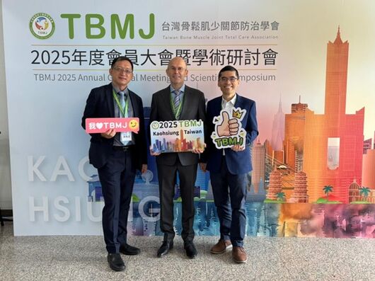 Auf dem Foto sind drei Personen abgebildet, die vor einer Leinwand stehen. Man erkennt im Hintergrund die Buchstaben TBMJ. Die drei Männer tragen Anzüge und schauen in die Kamera.