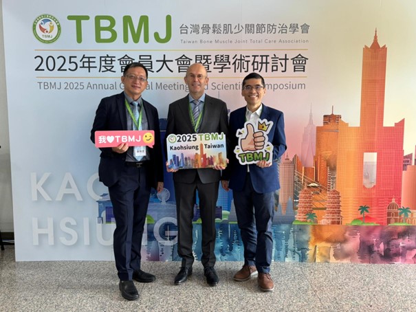 Auf dem Foto sind drei Personen abgebildet, die vor einer Leinwand stehen. Man erkennt im Hintergrund die Buchstaben TBMJ. Die drei Männer tragen Anzüge und schauen in die Kamera.