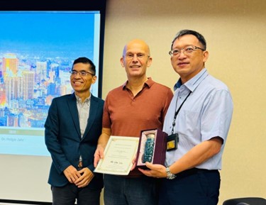 Auf dem Bild sind drei Männer zu erkennen. Der Mann links trägt einen Anzug und schaut nach links. Die anderen zwei Männer tragen Hemden und schauen zur Kamera. Der Mann in der Mitte hält eine Urkunde.