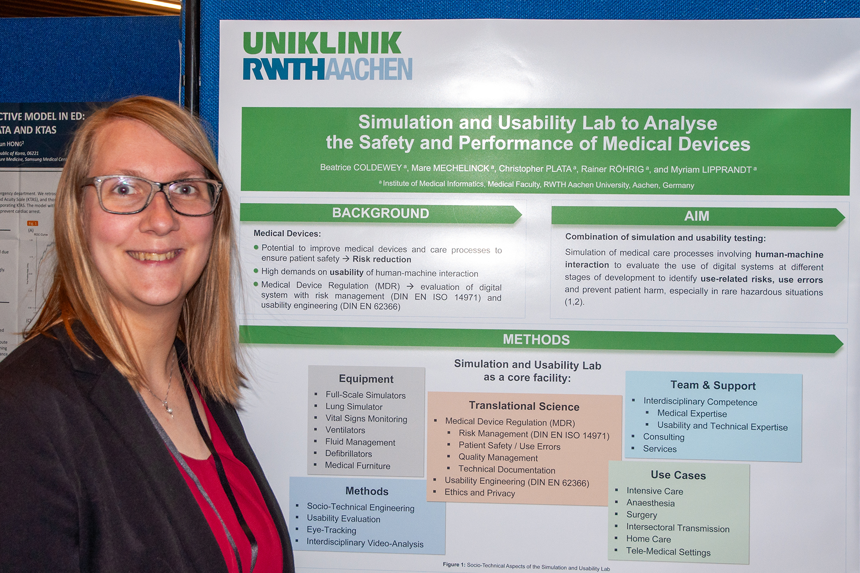 Links im Bild ist eine Frau mit langen rötlichen Haaren und Brille in einem roten Oberteil und schwarzer Strickjacke zu sehen, die neben einem wissenschaftlichen Poster steht. Das Poster ist im rechten Teil des Bildes gut erkennbar. Man kann die Überschrift "Simulation and Usability Lab to Analyse the Safety and Performance of Medical Devices" erkennen.