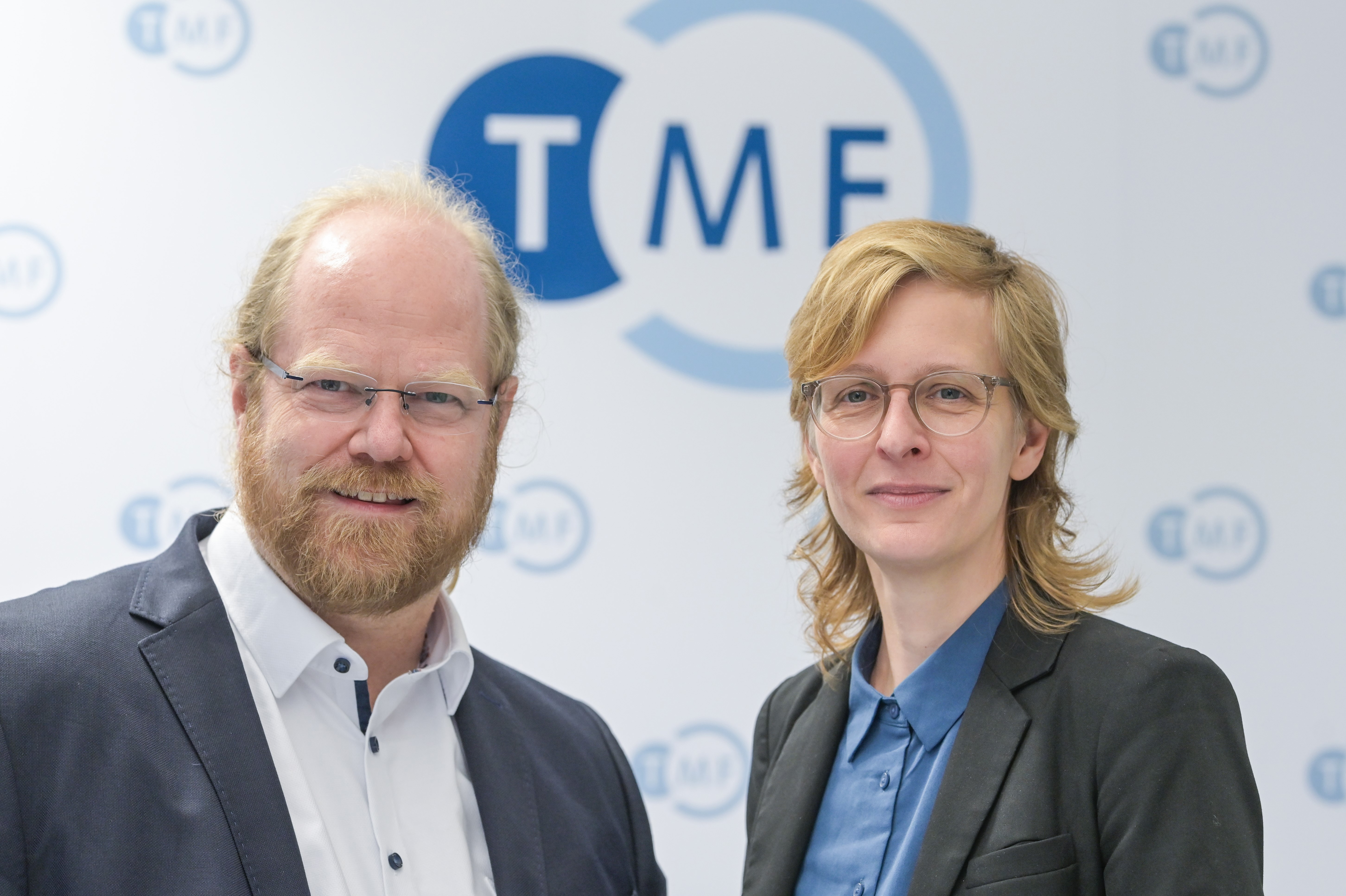 Bild von einem Mann und einer Frau vor einer Messewand mit dem TMF Logo. Beide Tragen Businesskleidung. Der Mann hat kurze rötliche Haare und einen Bart, die Frau trägt eine Brille und hat blonde schulterlange Haare.