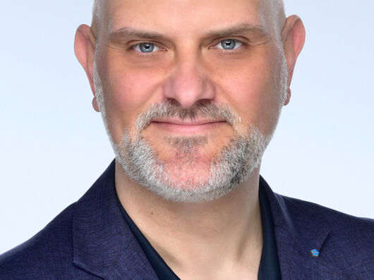 Ein mittelalter, weißer Mann mit grauem Bart und blauen Augen. Er trägt ein blaues Sakko.