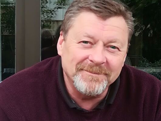 Ein älterer Herr mit angegrauten Haaren und Bart. Er trägt einen purpurnen Pullover.