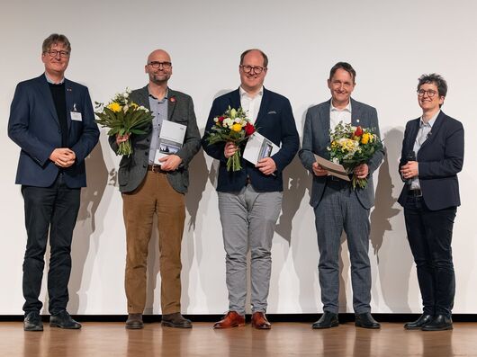 Bild der drei männlichen Preisträger mit Blumen und Urkunden. Ein Mann steht links und eine Frau rechts von ihnen