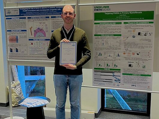 Mann hält eine Urkunde und steht vor zwei wissenschaftlichen Postern.