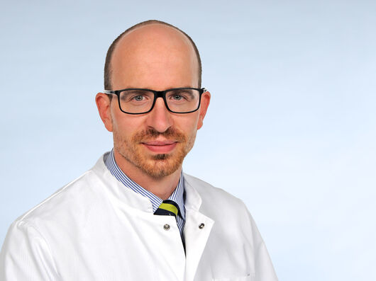 Porträtfoto eines Mannes mit hoher Stirn in einem weißen Arztkittel. Er trägt eine Brille und lächelt freundlich in die Kamera.