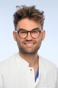Ein junger Mann mit gepflegtem 3-Tage-Bart und dunkler Brille lächelt nett in die Kamera