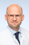 Porträtfoto eines Mannes mit runder Brille und kurzem Bart, der einen weißen Kittel trägt und freundlich in die Kamera lächelt.