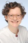Dr. med. Miriam Jansen Frau im weißen Kittel mit Brille und lockigen Haaren, die lächelt