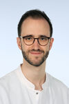 Portraitfoto eines Mannes mit dunklen Haaren, dunklem Bart und Brille. Er trägt einen weißen Arztkittel.