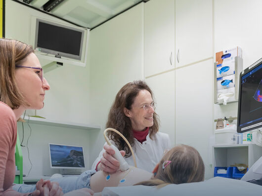 Das Bild zeigt eine medizinische Umgebung, in der eine Frau in einem weißen Arztkittel einen kleinen Patienten mit dem Ultraschallgerät untersucht.