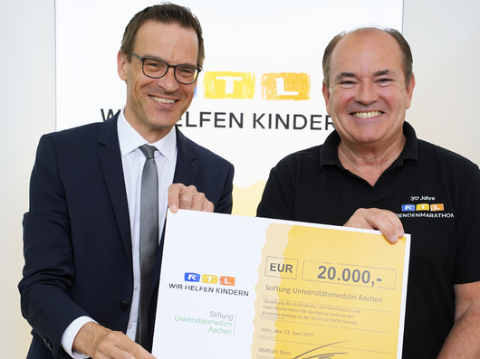 Ein Mann im Anzug mit Brille und ein älterer Mann in einem T-Shirt mit der Aufschrift RTL halten einen Scheck und lächeln in die Kamera