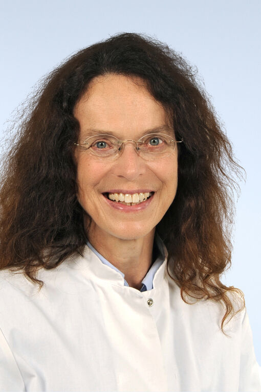 Porträtfoto einer Frau mit braunem Haar und durchsichtiger Brille, die einen weißen Kittel trägt und freundlich in die Kamera schaut.