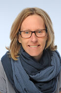 Portraitfoto einer Frau mit schulterlangen blonden Haaren und Brille in einem grauen Oberteil und einem blauen Schal, die freundlich in die Kamera lächelt.