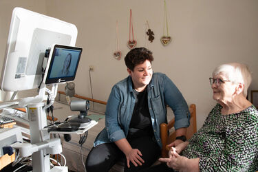 Eine Seniorin sitzt vor einem Bildschirm, auf dem eine weibliche Pflegeperson zu sehen ist, welche mit der Seniorin spricht. Neben der Seniorin sitzt eine Frau mit kurzen braunen Haaren und lächelt.