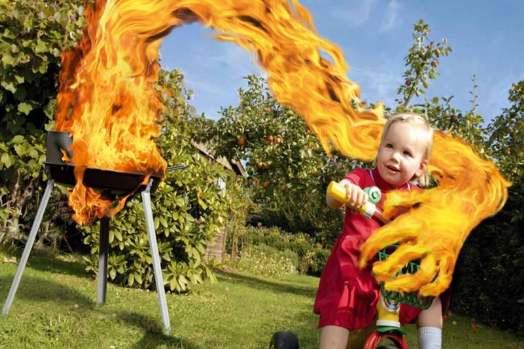 Bild eines Kleinkindes auf einem Dreirad vor einem brennenden Grill. Aus dem Grill greift eine Hand aus Flammen nach dem Kind.
