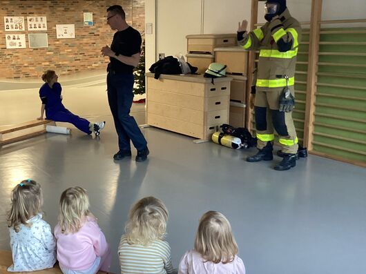 Kinder sitzen auf einer Holzbank und hören den Erklärungen eines Feuerwehrmanns zu