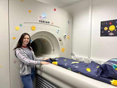 Das Bild zeigt eine Person, die neben einem MRT-Gerät steht, das mit verschiedenen Aufklebern von Sternen, Planeten und anderen Weltraumelementen dekoriert ist. An der Wand rechts vom MRT-Gerät ist ein weltraumthematisches Bild zu sehen. Die Person trägt legere Kleidung und hat eine Hand auf dem Bett des MRT-Geräts.