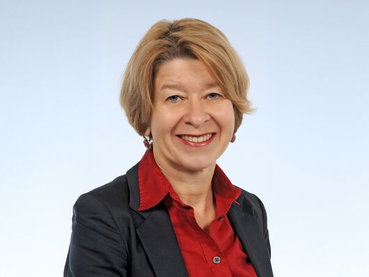 Porträt einer lächelnden Frau mit blonden Haaren. Sie trägt eine rote Bluse und ein dunkles Sakko.