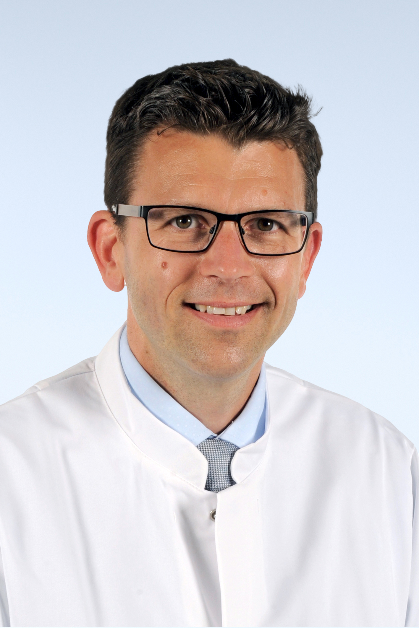 Porträtfoto eines Mannes mit kurzen, dunklen Haaren und einer Brille mit dunklem Brillengestell. Er trägt ein hellblaues Hemd und einen weißen Arztkittel. Er lächelt freundlich in die Kamera.