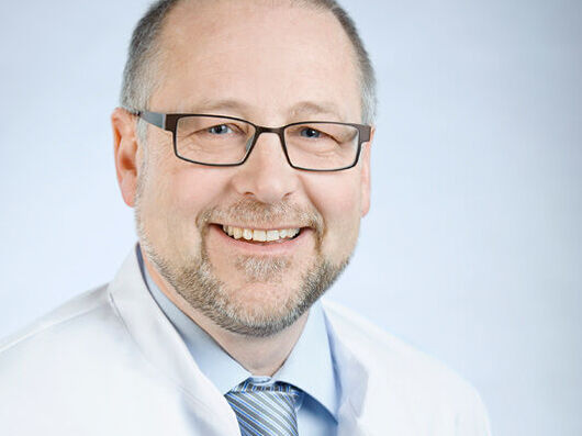 apl. Prof. Dr. rer. nat. Albrecht Eisert Porträtfoto eines Mannes mit hellen Haaren, Bart und Brille, der einen weißen Kittel trägt und freundlich in die Kamera lächelt.